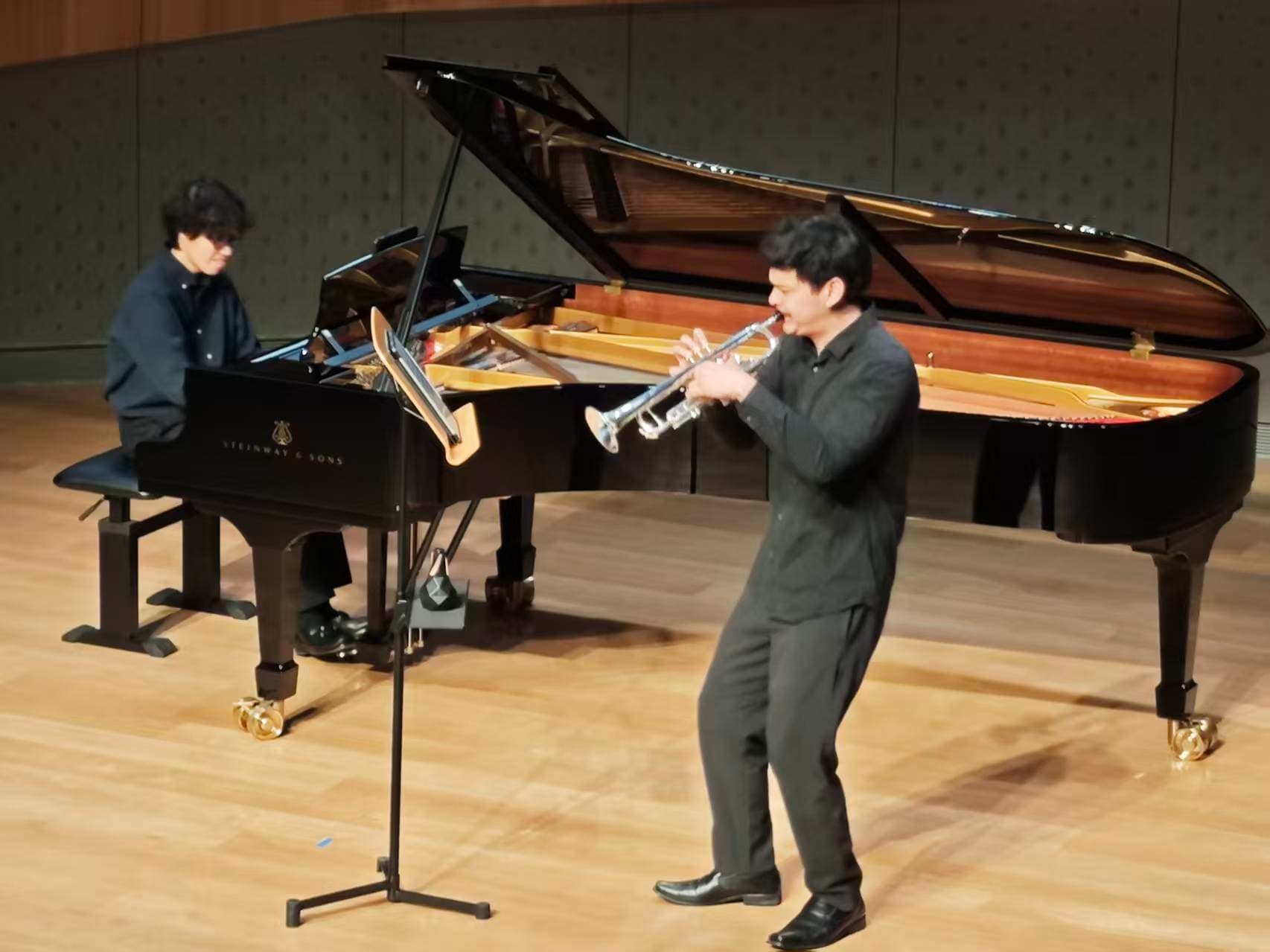 A trumpet performer playing beside a grand piano on a concert stage.